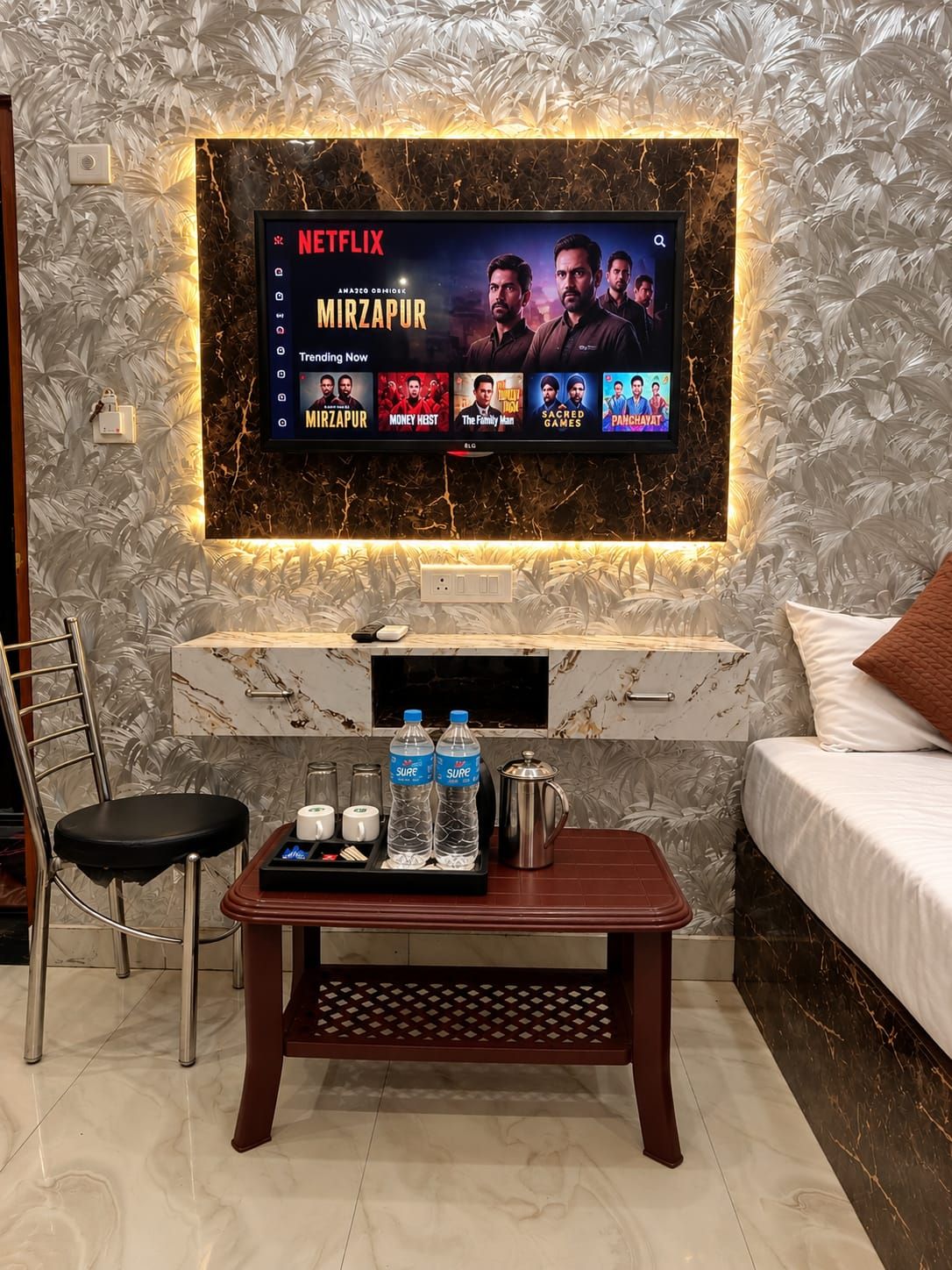 A modern hotel room with a large flat-screen TV mounted on a wall displaying a Netflix show, a wooden coffee table with water bottles and a coffee pot, a comfortable bed, and a chair.
