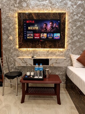 Une chambre d’hôtel moderne équipée d’un grand téléviseur à écran plat fixé au mur, sur lequel est diffusée une émission de Netflix, d’une table basse en bois avec des bouteilles d’eau et une cafetière, d’un lit confortable et d’un fauteuil.
