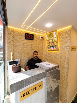 A reception desk with a 'RECEPTION' sign, a digital clock, and a decorative wall piece. - Trinetra Homestay (Varanasi)