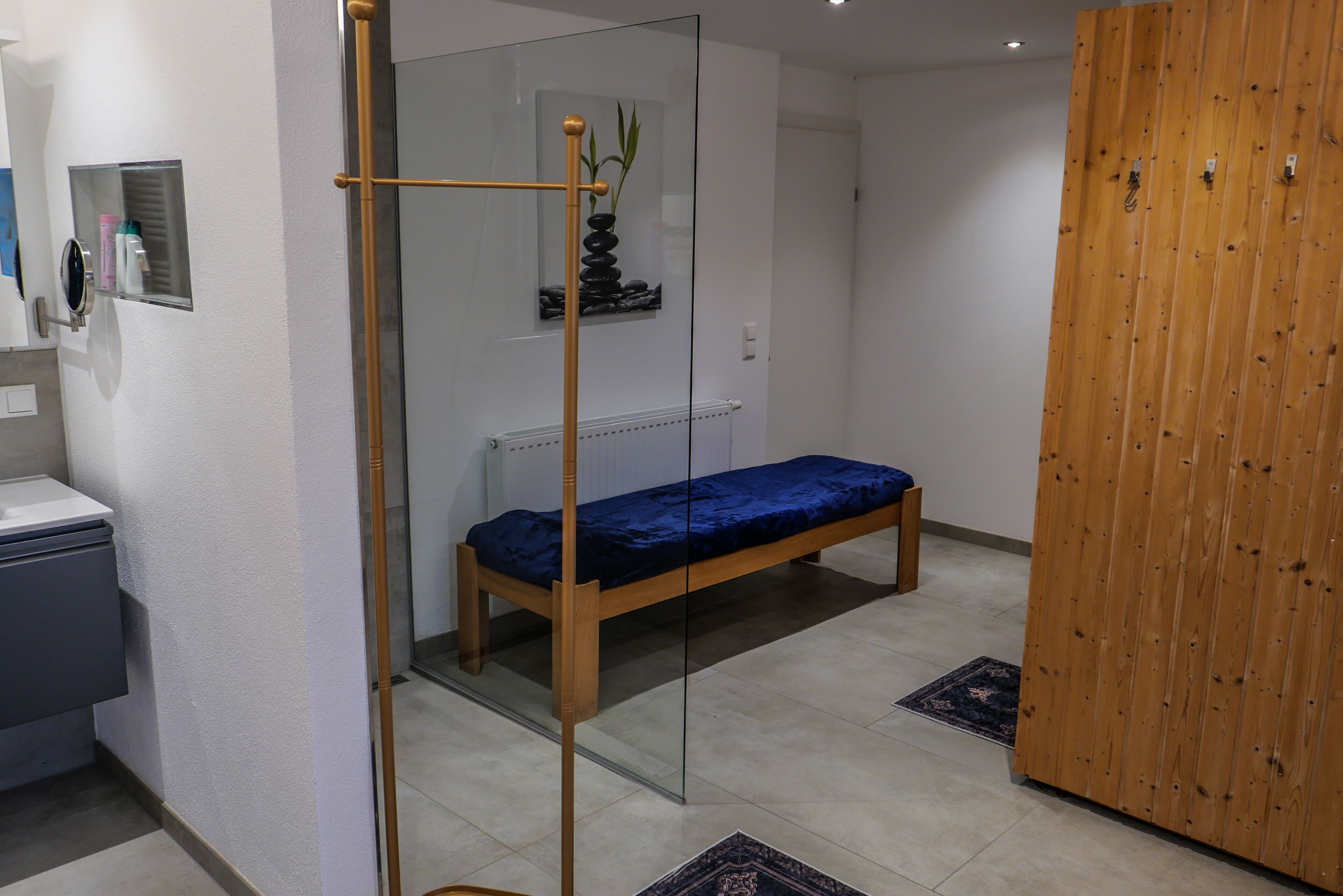 Une chambre avec un lit en bois, une cloison en verre, une petite étagère contenant des articles de toilette et une armoire en bois.