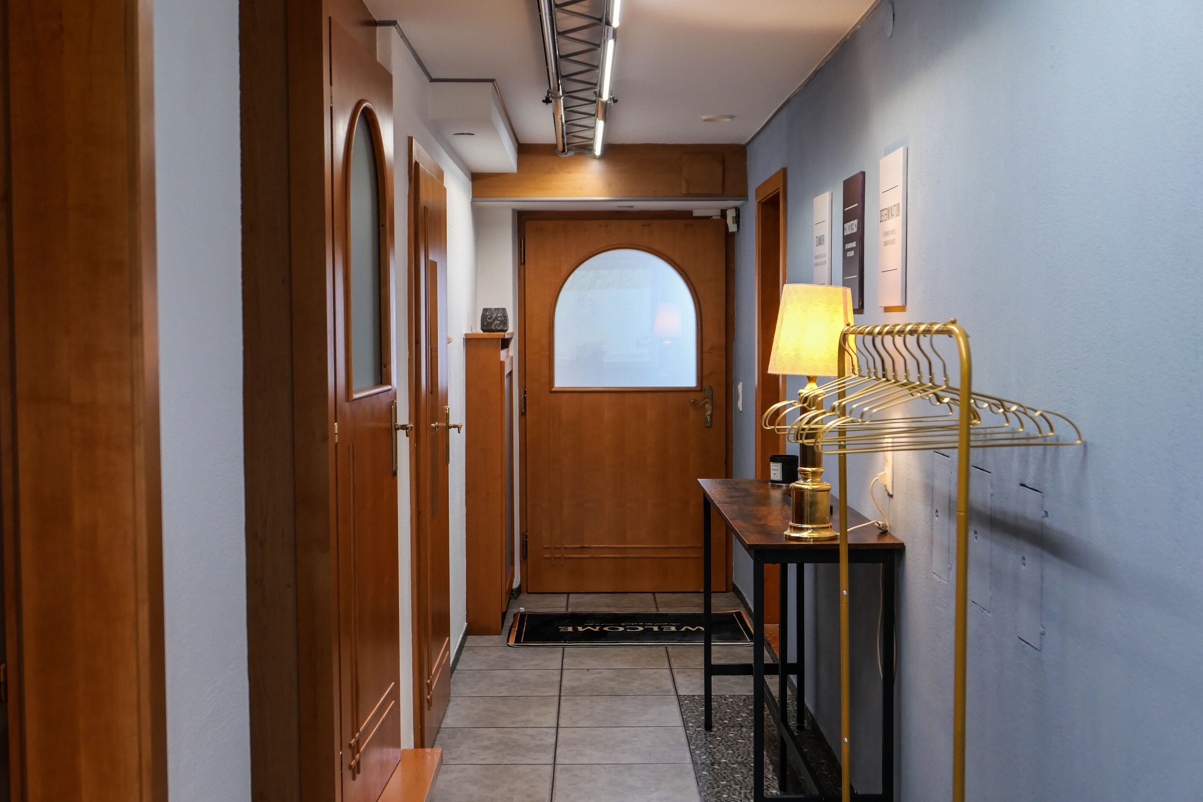 A hallway with wooden doors, a welcome mat, a lamp, and a metal rack.