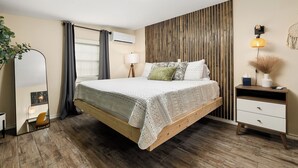 Een slaapkamer met een houten bed, een nachtkastje met een lamp, een spiegel en een wandairconditioner.