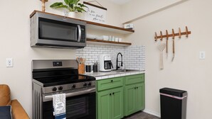 Een keuken met een magnetron, fornuis en groene kastjes. Er staat een koffiezetapparaat en een prullenbak.