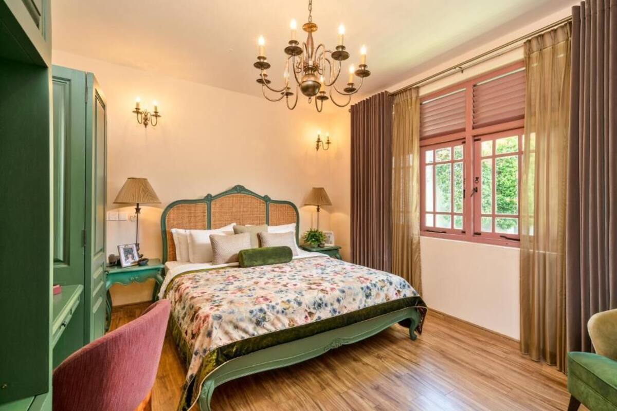 Un dormitorio con cama, candelabro, ventana con cortinas y mesita de noche con lámpara.