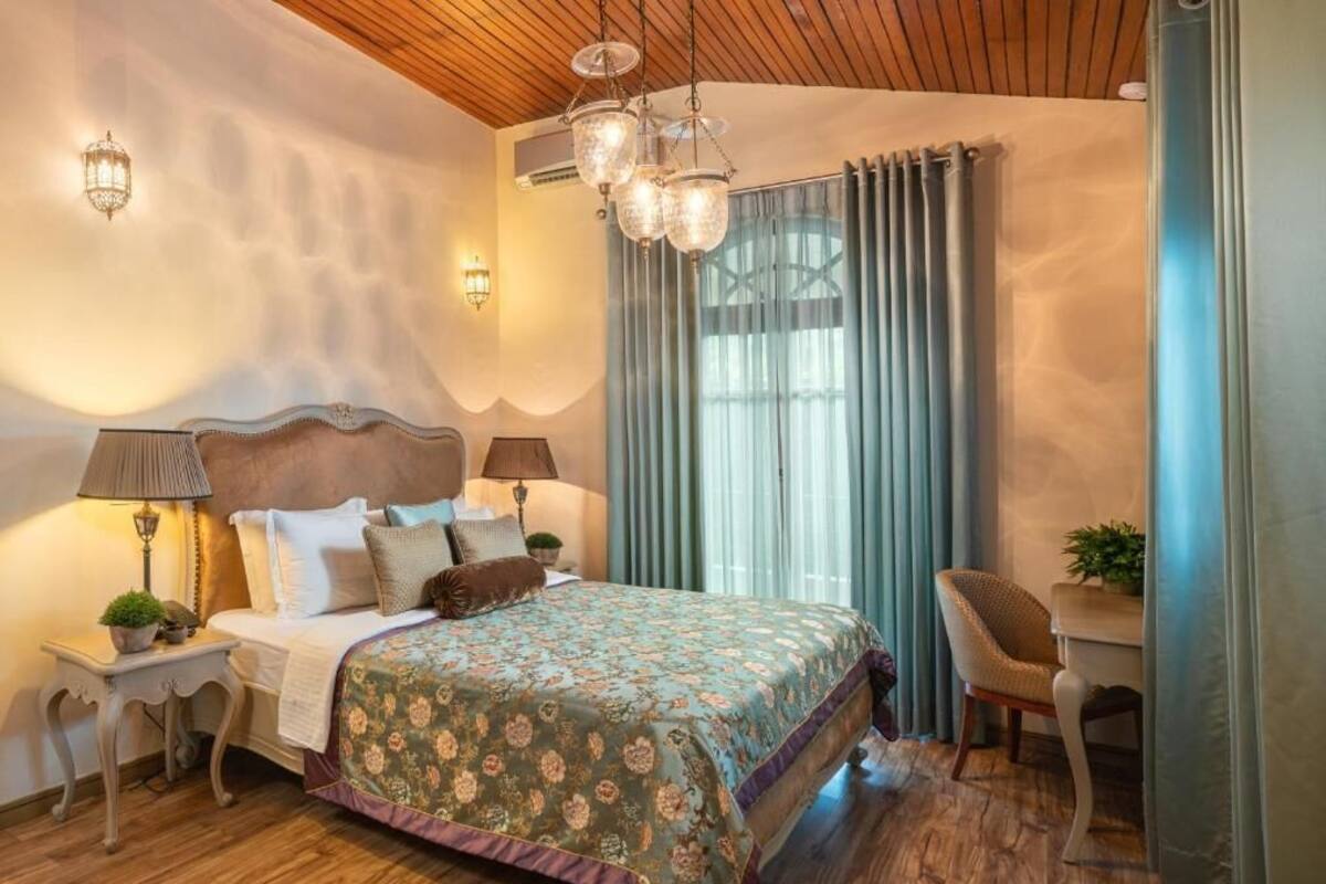 Un dormitorio con techo de madera, una cama grande con ropa de cama floral, dos lámparas de noche, una mesita con una silla y una ventana con cortinas.