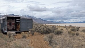 A trailer with a patterned interior, a gravel path, sparse shrubs, and a mountainous backdrop. - Dark Sky · Stargazer Horse Trailer · Edge of the Alvord Desert (Princeton)