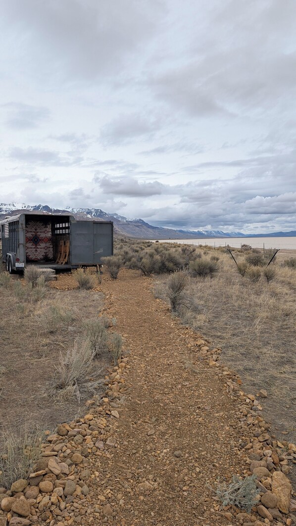 Una caravana con un interior estampado, un camino de grava, arbustos dispersos y montañas al fondo.