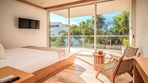 Una camera da letto con un letto grande, una TV fissata al muro, un balcone vista alberi e una casa, e sedie in vimini.