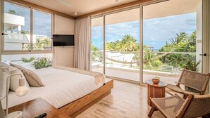 A modern bedroom with a large bed, a TV mounted on the wall, a balcony with a view of palm trees and buildings, and wicker furniture. - Oceanfront Apartment in Greta – Luxury & Style (Puerto Morelos)