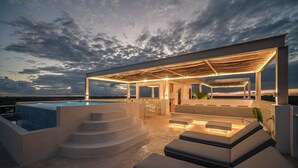 A modern outdoor pool area with a covered seating area, steps leading to a pool, and a view of the ocean at dusk. - Rooftop Pool, Fast WiFi & Nature Views at Greta (Puerto Morelos)