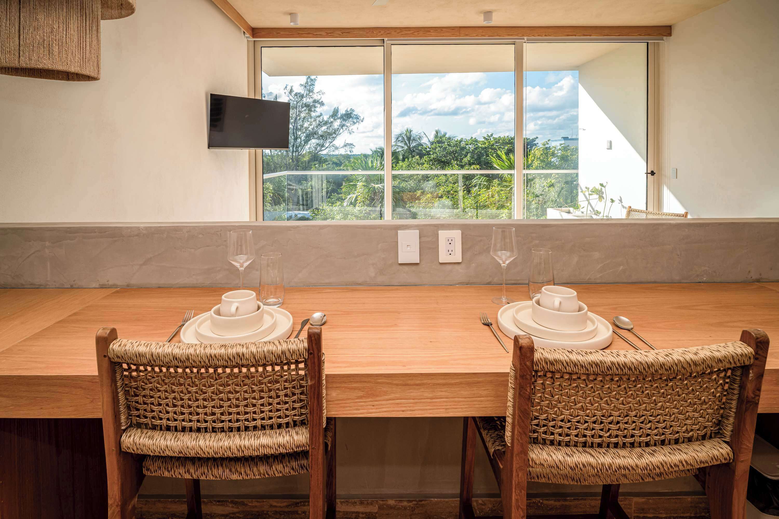 A dining area with a long wooden table, wicker chairs, and a large window offering a view of greenery and a building.