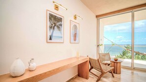 A modern room with a wooden desk, two framed pictures, a white vase, and a wicker chair. - Modern Seafront Unit | Infinity Pool, Gym (Puerto Morelos)