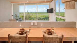 A dining area with a long wooden table, two wicker chairs, and a view of a green landscape through large windows. - Modern & Peaceful Getaway | Eco-Luxury in Greta (Puerto Morelos)