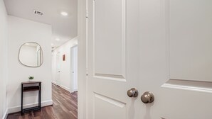 A hallway with a wooden floor, a white door with a silver handle, a small table with a plant, and a round mirror on the wall. - Houston Hideaway Steps from Rice Village Fun (Houston)