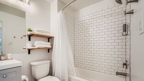A modern bathroom with a white toilet, a bathtub, and a shower with a white curtain. There are shelves above the toilet holding towels and toiletries. - Houston Hideaway Steps from Rice Village Fun (Houston)