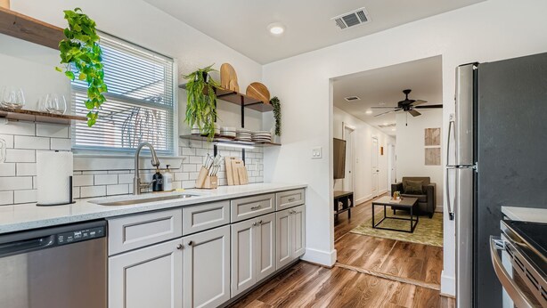 A modern kitchen with white cabinetry, a dishwasher, and a refrigerator. There is a window with blinds, a ceiling fan, and a dining area with a table and chairs. - Houston Hideaway Steps from Rice Village Fun (Houston)