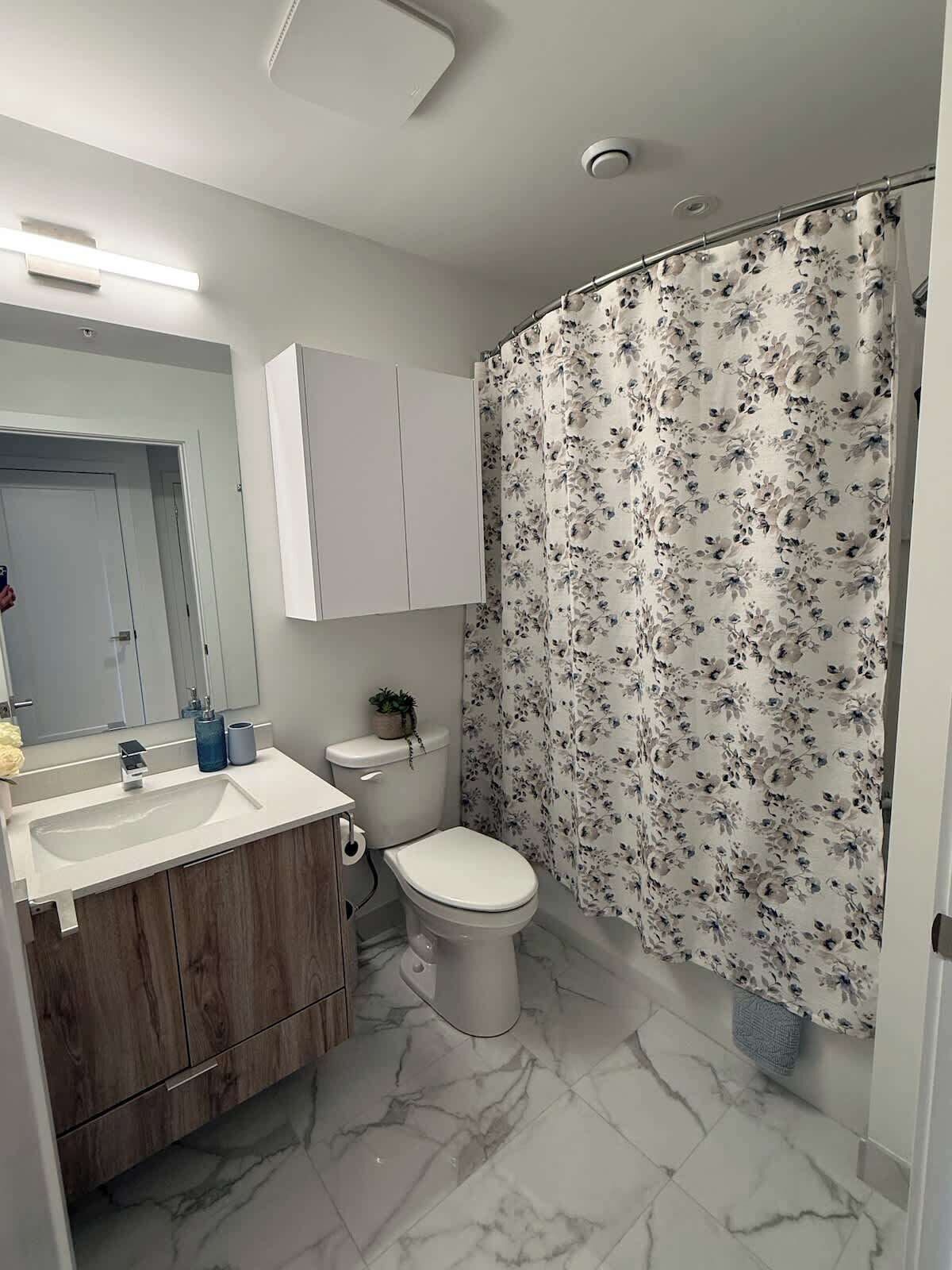 Une salle de bain moderne avec un sol en marbre, un lavabo blanc, des toilettes et un rideau de douche à motifs floraux.