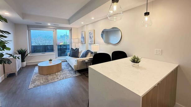 Una sala de estar moderna con sofá, mesa de centro y zona de comedor. Hay un ventanal con vistas a edificios.