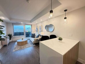 Sala moderna com sofá, mesa de centro e área de jantar. Há uma janela grande com vista para edifícios.