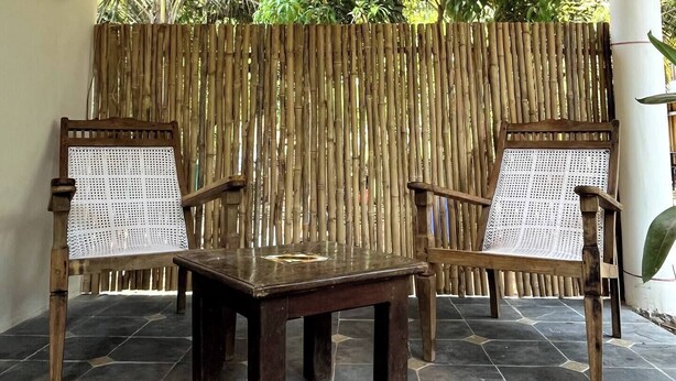 Uma área de estar coberta ao ar livre, com duas cadeiras de madeira e uma mesa, parede de bambu e telhado de palha.