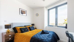 A bedroom with a bed, a nightstand, a lamp, a clock, a window, and a chair. - Luxury Apt Near Canary Wharf (Greater London)