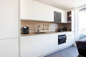 A modern kitchen with white cabinetry, a wood backsplash, a coffee machine, a sink, and a stove. - Luxury Apt Near Canary Wharf (Greater London)