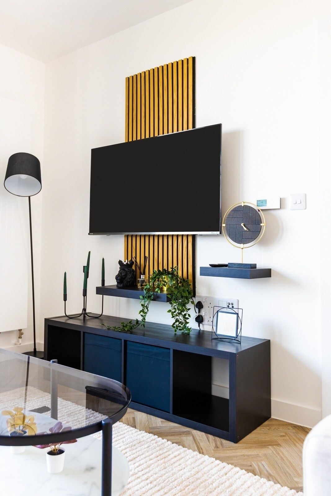 A modern living room with a flat-screen TV mounted on a wooden panel, a black lamp, a round clock, and a dark-colored entertainment unit with decorative items.