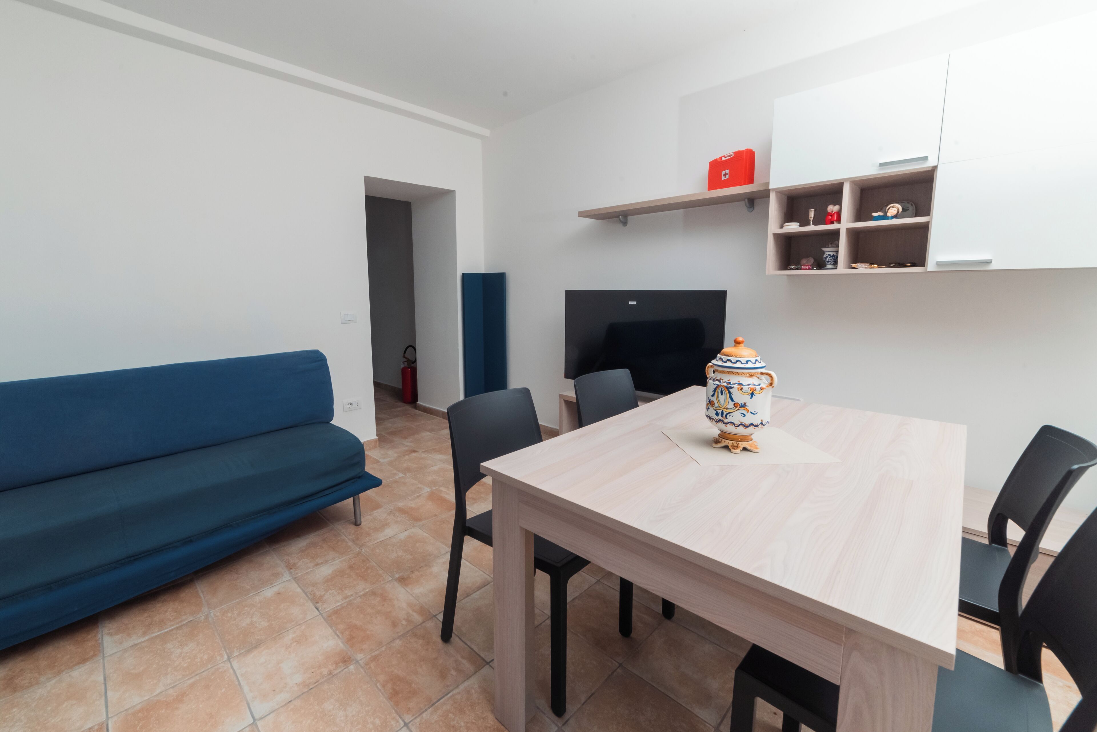 Una sala moderna con un sofá azul, una mesa de comedor de madera y sillas negras. Hay un televisor montado en la pared y un estante con varios objetos.