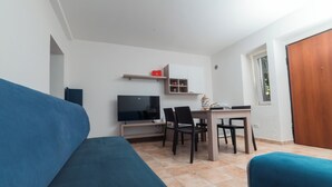 Een knus woongedeelte met een bank, een televisie, een eettafel met stoelen, en een klein kastje met planken.