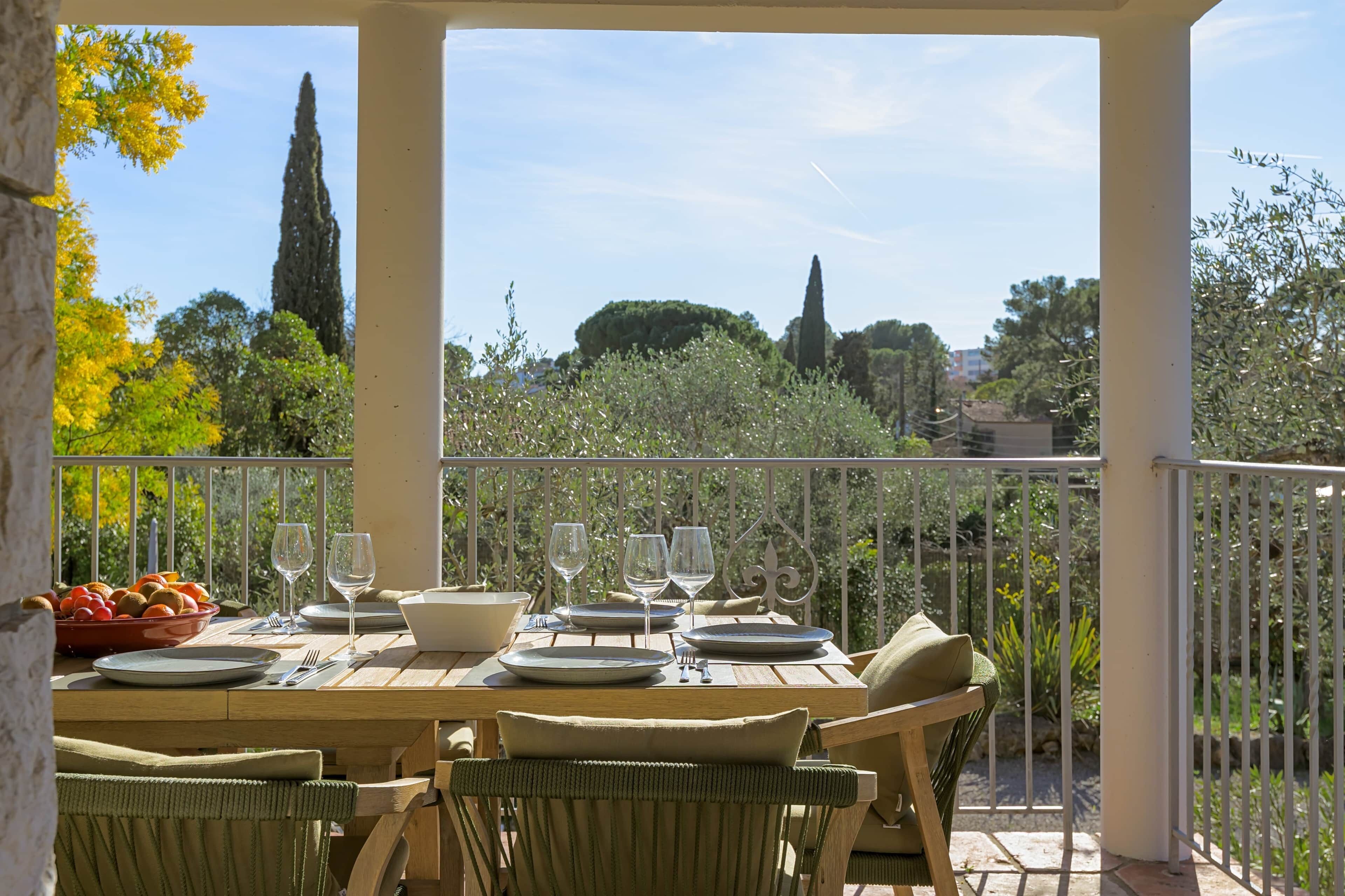 Ein Essbereich im Freien mit einem für vier Personen gedeckten Tisch, umgeben von Stühlen und Blick auf einen Garten mit Bäumen und blauem Himmel.