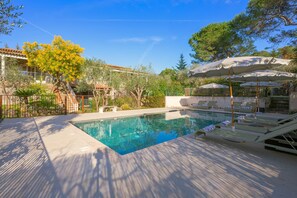 Une piscine entourée d’arbres et d’une clôture, avec des chaises longues et des parasols à proximité.