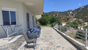 Une terrasse avec un sol en pierre, une table, des chaises et une balustrade donnant sur une colline parsemée de bâtiments.
