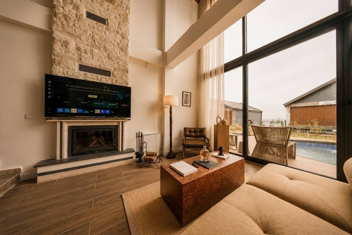 A living room with a stone fireplace, a flat-screen TV, a sofa, and a view of a pool and outdoor area.
