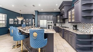 A modern kitchen with dark blue cabinetry, a marble island, and blue bar stools with gold accents. - Hollywood Glam Villa w/ AGT Stage Arcade Pool Spa! (Kissimmee)