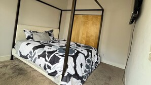 Una cama individual con estructura metálica negra y colcha de estampado floral en una habitación con vigas de madera a la vista en el techo.