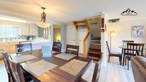 Una cocina moderna con armarios blancos, una mesa de comedor de madera con individuales, y una escalera que sube a una zona de altillo.