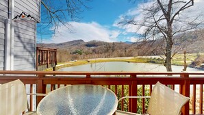 Una terraza de madera con una mesa y sillas de vidrio, con vistas a un estanque y montañas.