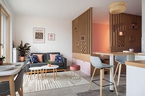 Una sala de estar moderna con sofá, mesa de centro y sillas. Hay un comedor con mesa y sillas. La habitación tiene paneles de madera y una lámpara colgante.
