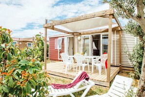 Eine Holzterrasse mit Gartenmöbeln, einem überdachten Sitzbereich und Blick auf ein rotes Gebäude.