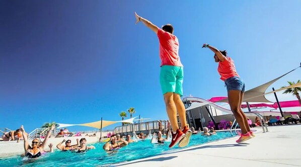 Ein Schwimmbecken mit Personen, die springen und schwimmen, umgeben von einer sonnigen Außenanlage.