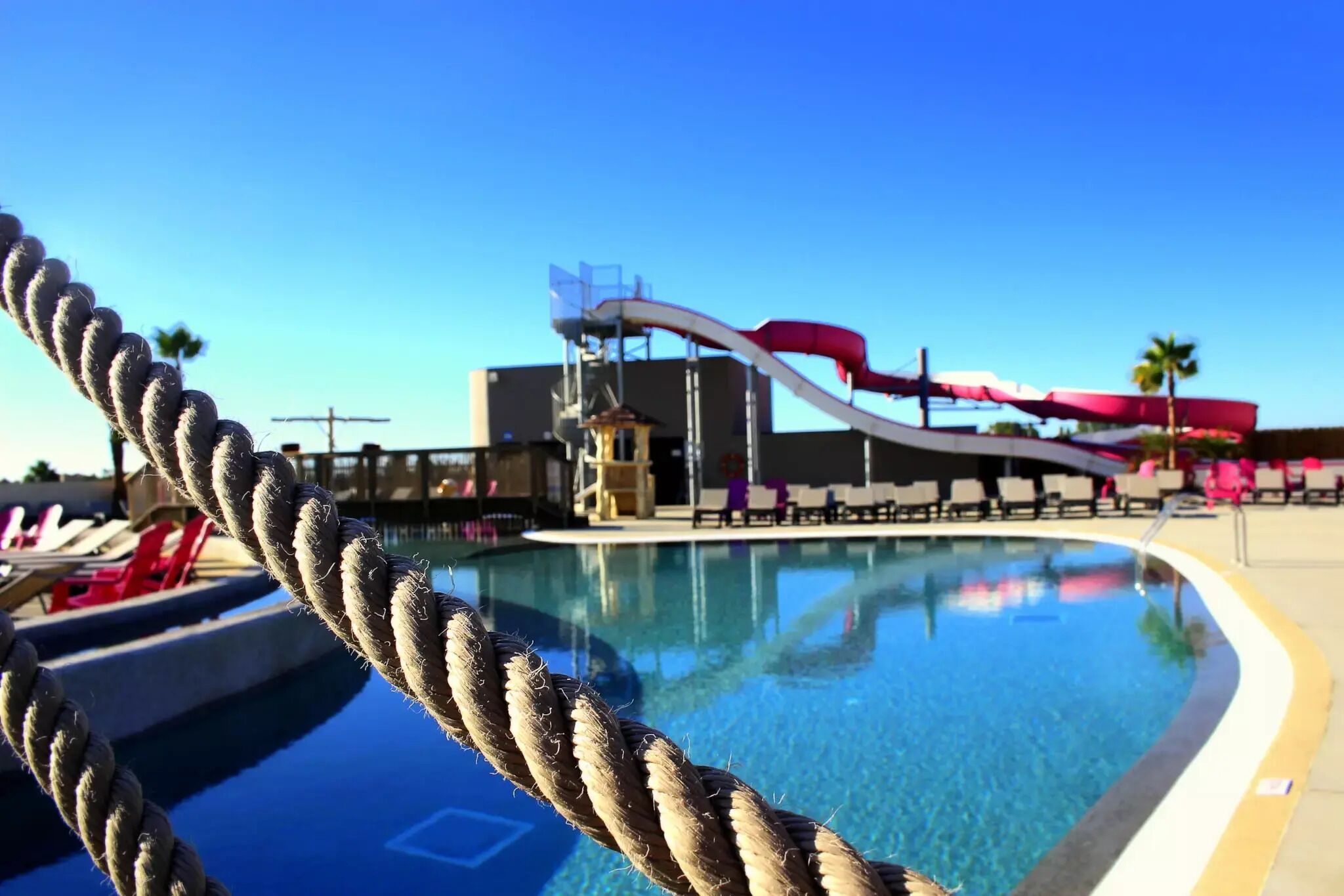 A pool with a rope barrier, a water slide, lounge chairs, and a building structure in the background.