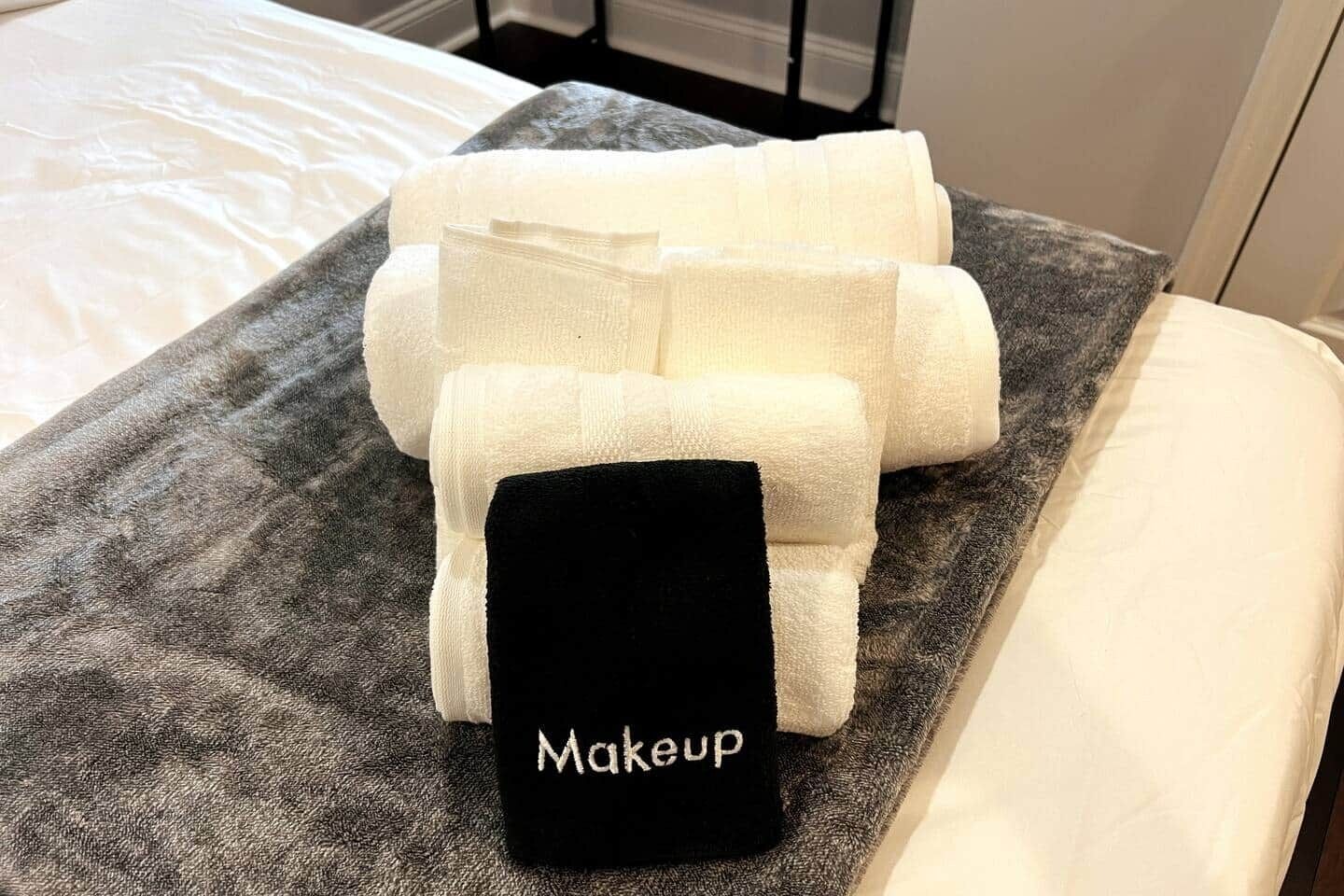 Cama com lençóis brancos e almofada preta com a etiqueta 'Maquiagem' sobre uma superfície cinza texturizada.