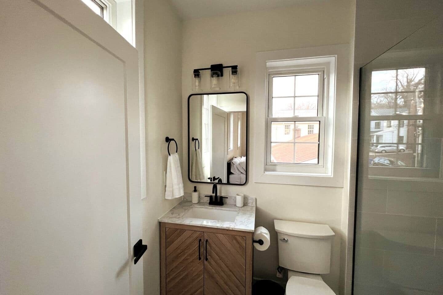 Un baño con tocador de madera, espejo, inodoro y ventana con vista a un edificio.