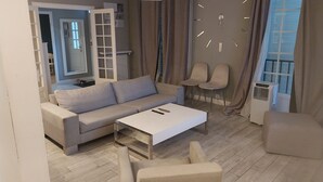 A modern living room with a sofa, a coffee table, and two chairs. There is a large window with curtains, a wall clock, and a ceiling light fixture. - Spacious and pleasant (Garges-lès-Gonesse)