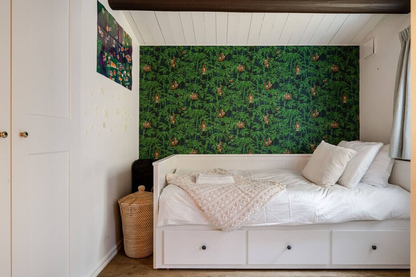 Une chambre avec un lit blanc, un panier en osier et un papier peint vert à motifs de feuilles.