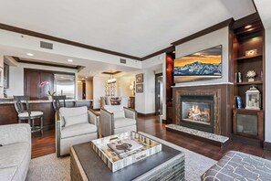 A modern living room with a fireplace, comfortable seating, a kitchen area, and a large TV mounted above the fireplace. - Three-Bedroom · 3.5 Bath · Top-Floor · Sleeps 8 (Truckee)