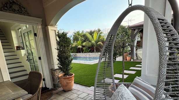 A hanging chair with cushions, a dining table, and chairs, a potted plant, and a view of a swimming pool and garden.
