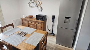 A dining area with a wooden table, chairs, a microwave, a refrigerator, and a wall-mounted light fixture. - Apparently (Naintré)