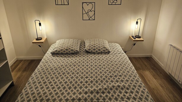 A bedroom with a patterned bedspread, two bedside tables with lamps, and three framed artworks on the wall. - Apparently (Naintré)