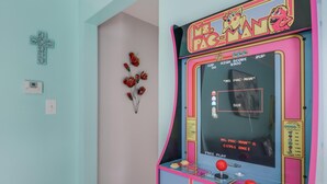 Une borne d’arcade Pac-Man vintage, dans une pièce aux murs bleu clair, ornée d’une croix décorative et agrémentée de fleurs séchées.